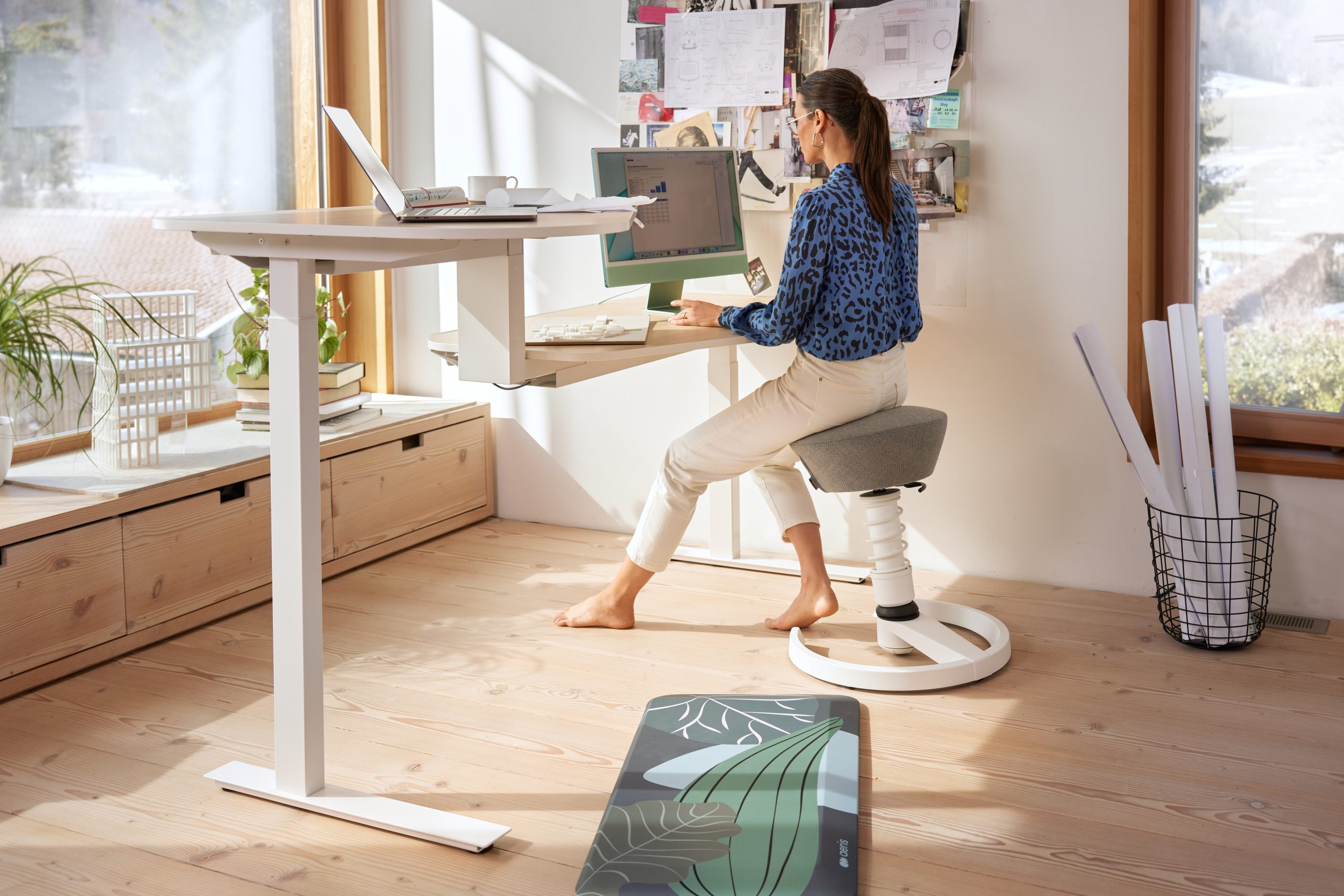 Woman sits at a height-adjustable desk typing on a laptop in a bright home office with a corkboard wall and plants nearby.