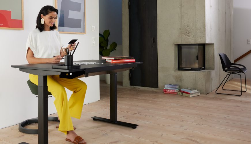 Woman at a height-adjustable desk, looking at photos, in a bright modern room with framed abstract art on the wall, left side of the image.
