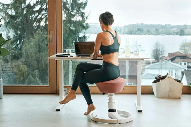 Woman in activewear sits on a circular balance stool at a desk with a laptop, by a large window overlooking a lake.